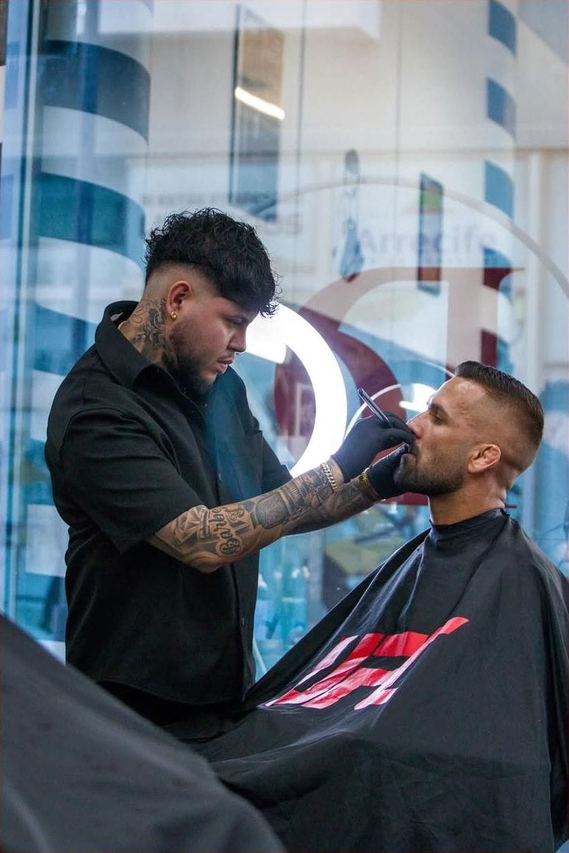 Barbero arregla la barba de un cliente con capa de la UFC junto a un aro de luz