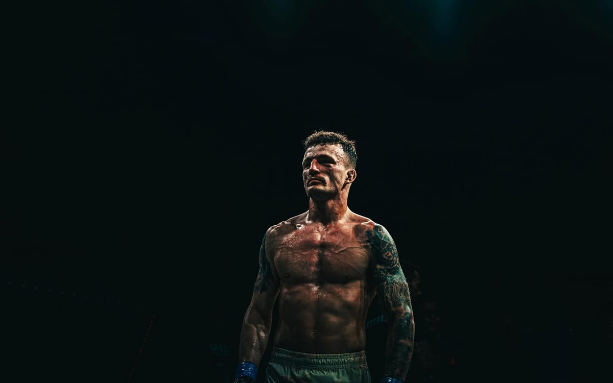 Peleador con los brazos tatuados de pie en la oscuridad de la jaula, iluminado desde arriba tras su combate