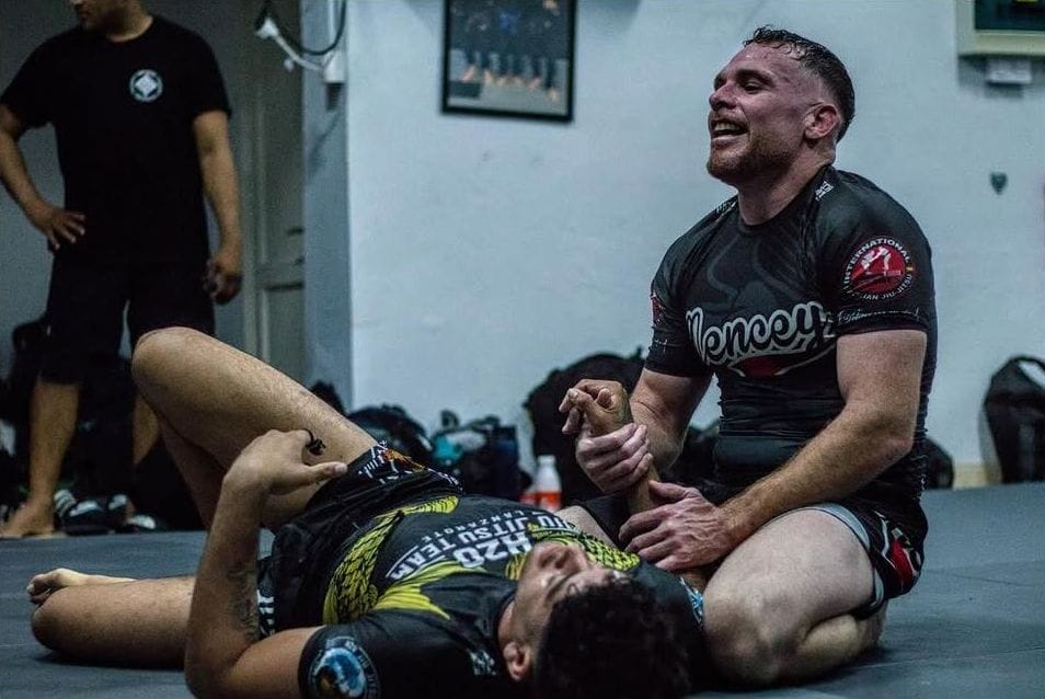 Entrenador sonríe mientras controla a un compañero en el tatami, con el resto del grupo detrás