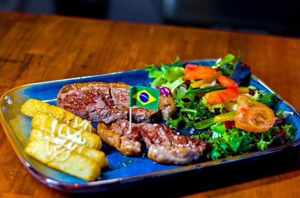 Plato de carne a la brasa con ensalada, yuca y una bandera de Brasil