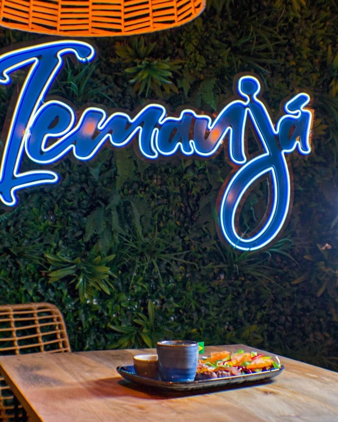 Rótulo de neón azul de Iemanjá sobre un muro vegetal, con un plato servido en la mesa