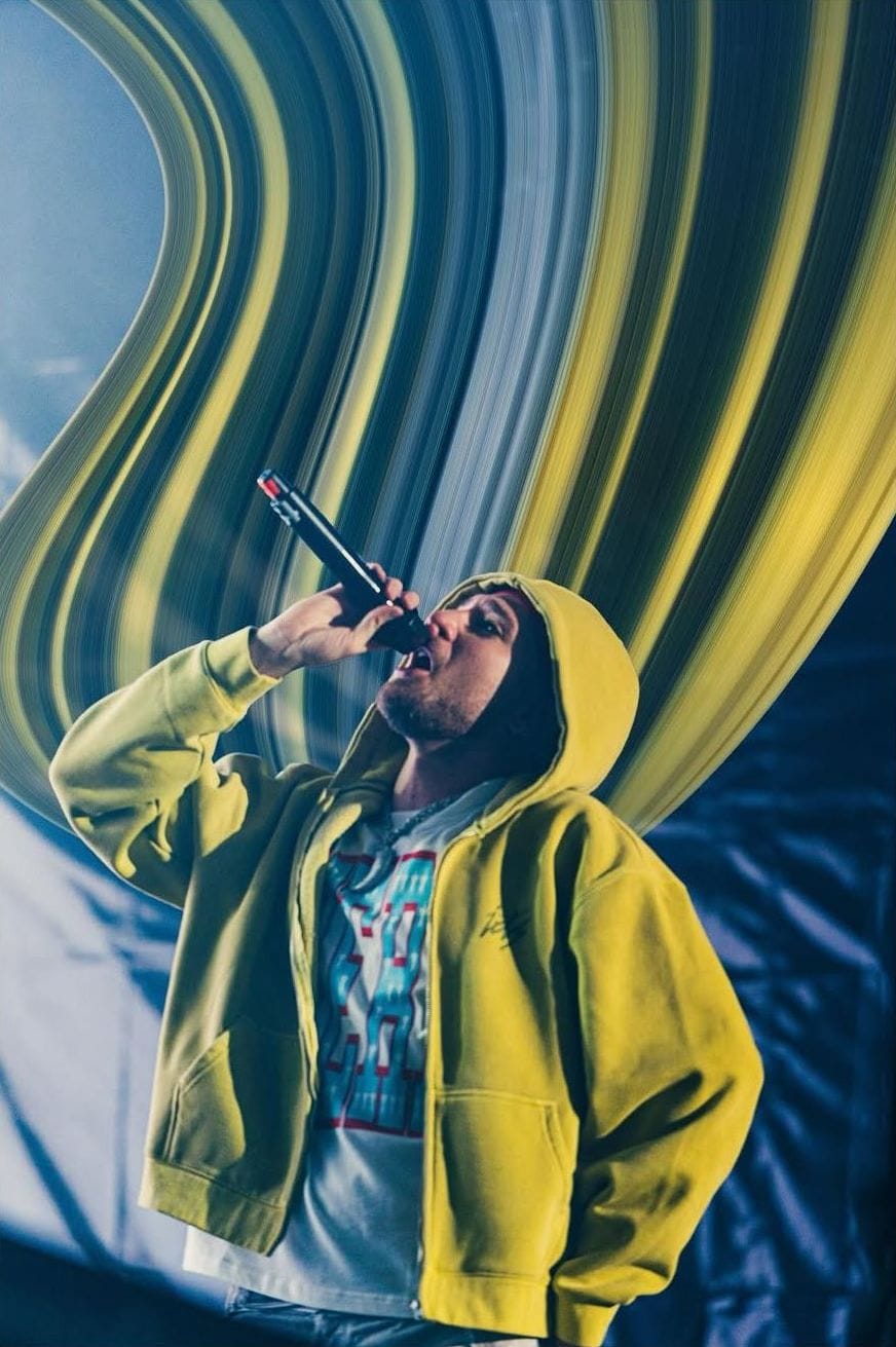 Artista con sudadera amarilla canta con el micrófono en alto entre franjas de color estiradas
