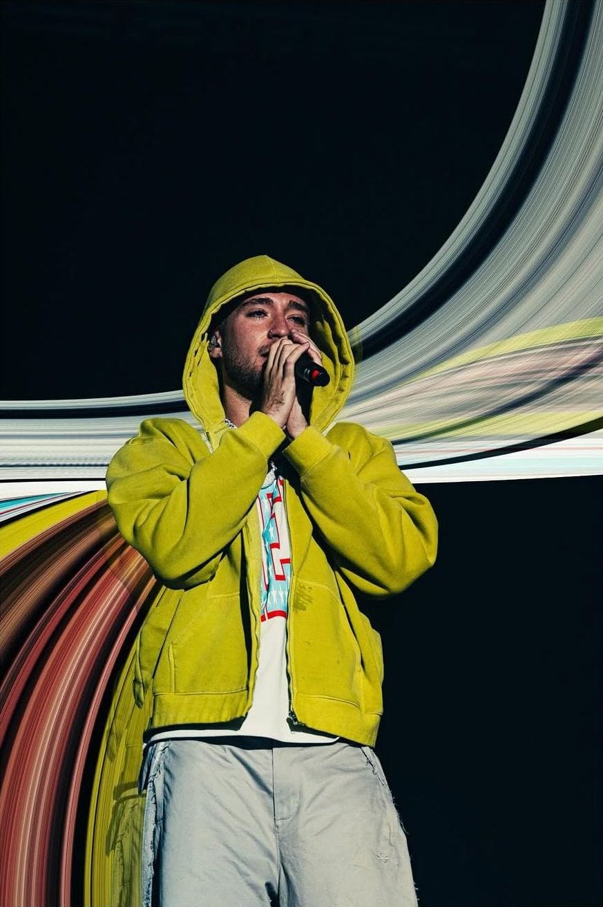 Artista con sudadera amarilla canta con las manos juntas entre ondas de color
